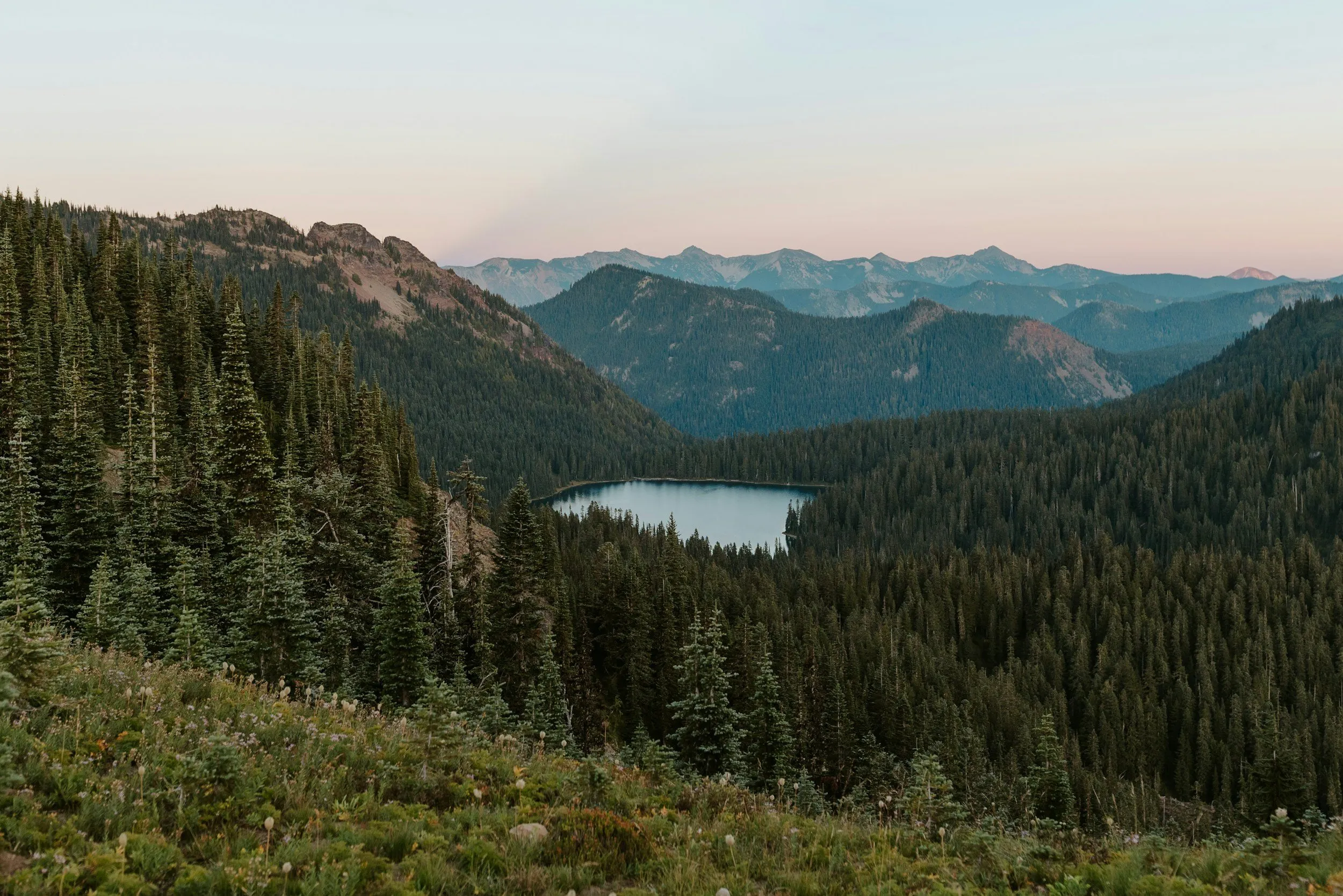A lake surrounded by evergreen forest, with layered mountain ridges beyond.
