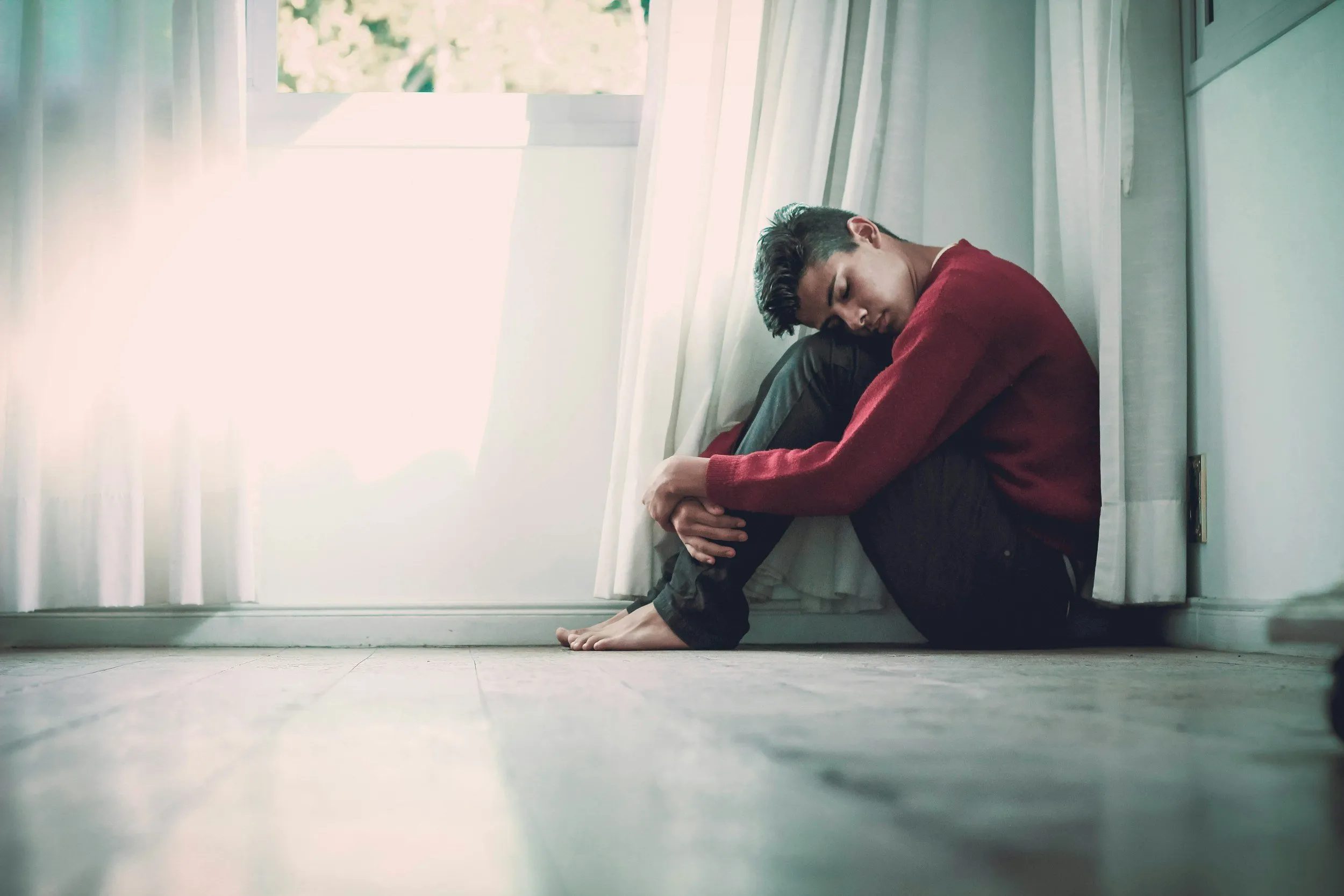 A person in a red sweater sitting on the floor beside a window, with their head resting on their knees.